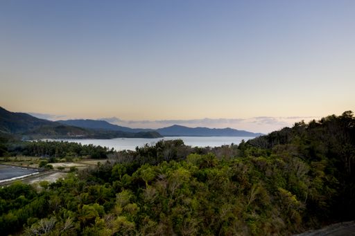 A high angle shot of forested hills and shore near the sea with mountains in the distance under a blue sky