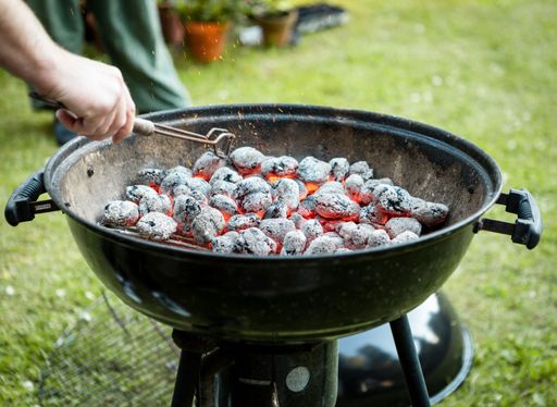 A closeup of person's hand with metal tool turning around burning charcoal in black barbecue in the yard