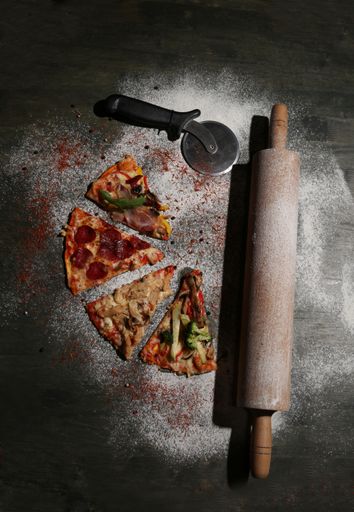 A top view of a cut pizza with a rolling pin ant a pizza cutter with flour on a wooden table