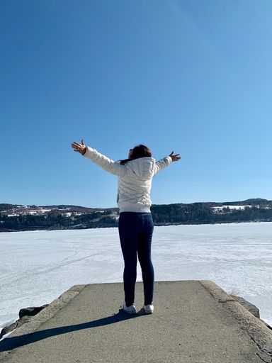 A vertical shot of a cheerful female standing on the edge of a road leading into a frozen lake with her hands open against a blue sky