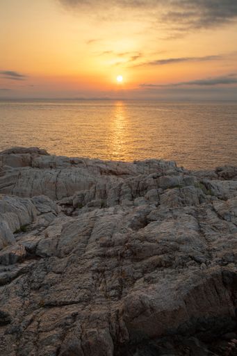 A vertical shot above a rocky cliff and the setting sun coloring the surface of the tranquil sea
