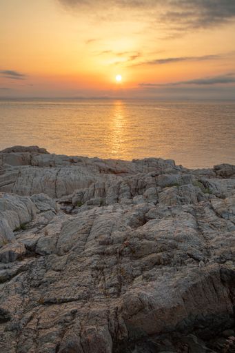 A vertical shot of a rocky cliff and the reflection of the setting sun on the sea