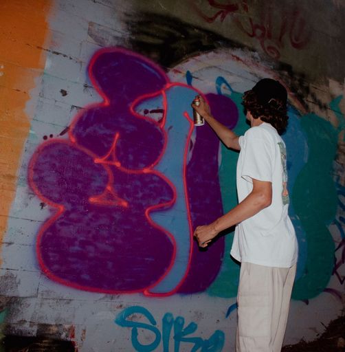 A teenager in a baggy white t-shirt spray painting graffiti under a bridge in Santa Cruz, California