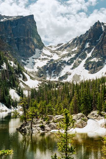 A gorgeous vertical shot of tall snowy mountains overlooking lush green trees near a lake