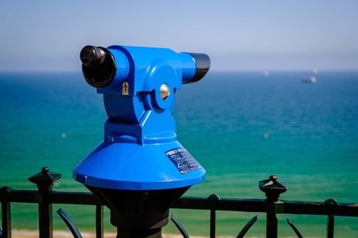 A blue public telescope on the shore, for touristic usage, with a turquoise seascape in the background, on a sunny day