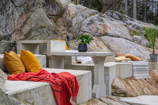 A concrete table and bench set outdoors on a rock with few palm plants and throw pillows