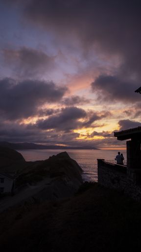 A vertical shot of a cottage surrounded by rocks and the sea under a cloudy sky during the sunset