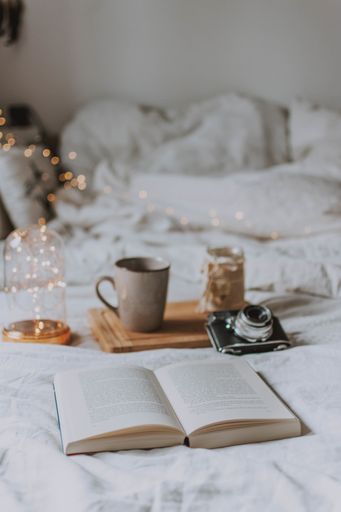A vertical shot of an open book, a camera, a tray and a mug on a bed with white sheets