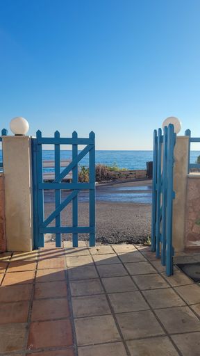 A vertical closeup shot of a blue wooden fence door leading to an ocean shore on a sunny day