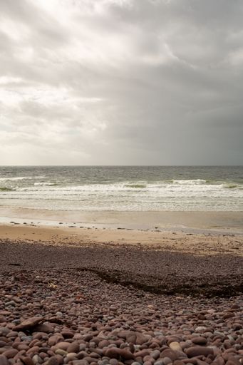 A vertical shot of a rocky beach with the foamy sea and the horizon in the background on a gloomy day