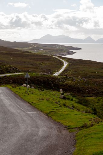 A vertical shot of a countryside road surrounded by fields, a lake with mountains in the background