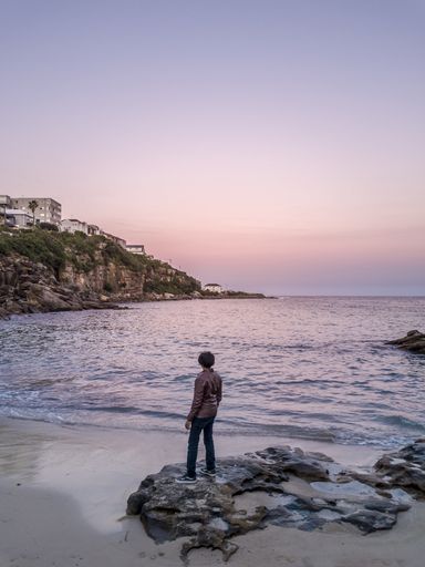 A male model in a coat standing on rocks at the shore of the sea enjoying the beautiful sunset
