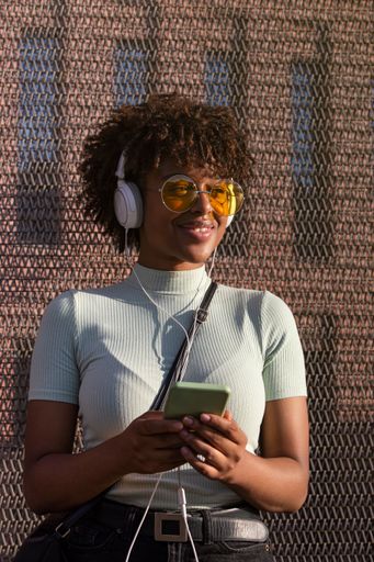 A cool young latin american woman with afro hair and glasses smiling listening to music on smartphone with headphones