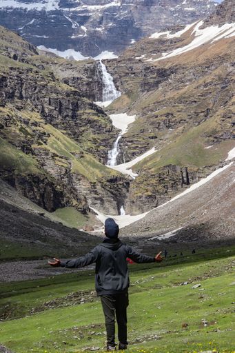 A rear view of a man standing against a beautiful view of a waterfall in mountains and enjoying it with arms opened