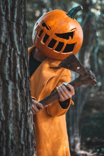 A vertical shot of a young woman wearing a pumpkin head and holding an axe