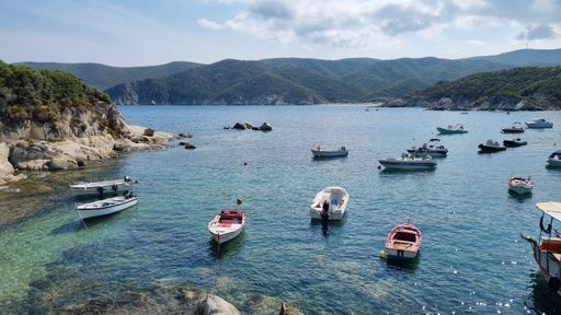 A seascape with boats in front of the mountains and a cloudy blue sky