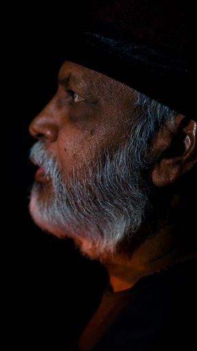 A vertical portrait of a South-Asian man with grey beard and a hat - looking thoughtful