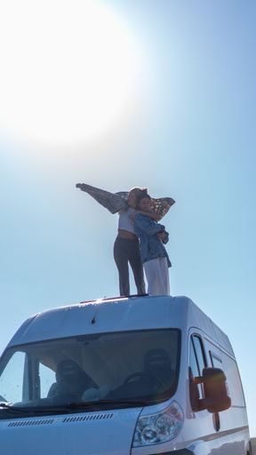 A vertical shot of two female friends smiling and hugging on top of a bus during a road trip