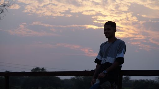 A South East Asian guy posing against a railing against a cloudy sky at sunset