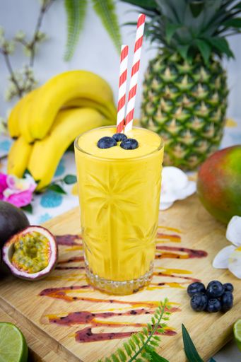 A vertical top view of a yellow drink with blueberries on it with tropical fruits on the board