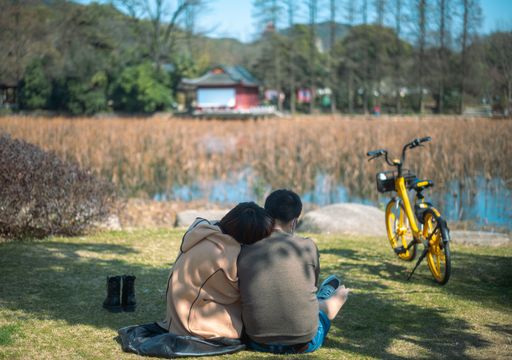 A couple sitting on grass park wth a bicycle looking at the lake with blur trees