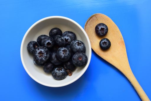 A high angle shot of a white bowl with blueberries near a wooden spoon isolated on a blue background