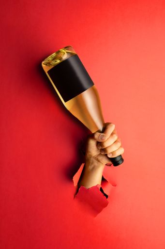 A human hand sticking out of torn hole in red paper holding wine bottle with blank label