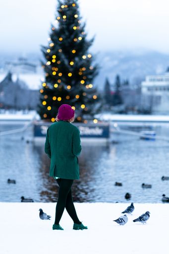 A vertical shot of a female in a coat standing with pigeons against a lake and a blurry Christmas tree