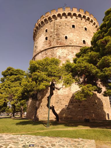 A vertical shot of the White Tower of Thessaloniki with trees under a clear blue sky
