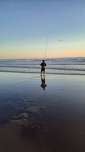 a single fishing man with a long rod at sunset time at Parangtritis Beach on wet and reflecting sand, Java, Indonesia