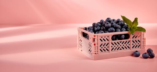 A pile of sweet blueberries and sprig of mint in plastic container placed on pink background