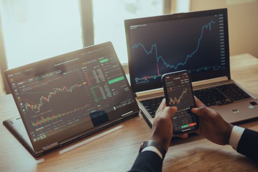 A back shot of a man holding a smartphone next to laptop trading online with chart crypto stocks