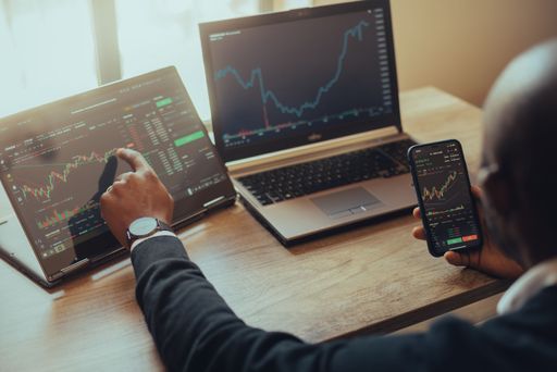 A young man holding a smartphone and laptop with online trading chart of crypto on the screen
