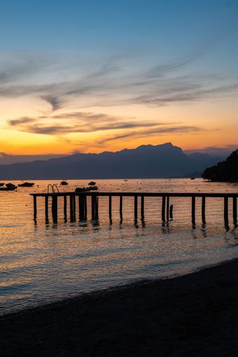 A vertical shot of a pier on the shore of Lake Garda at sunset with ships in the background, Lazise, Italy