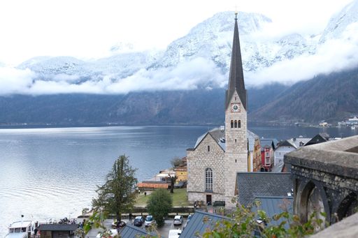 The tower of a church overlooking a calm lake surrounded by mountains with their peaks covered by clouds in Halstatt, Austria
