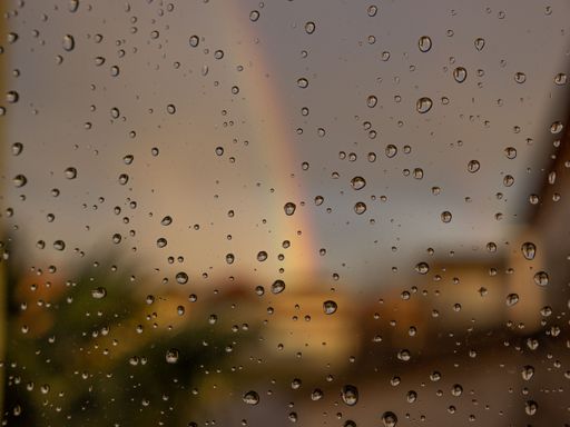 A closeup of raindrops on a glass window on a blurry background of a rainbow