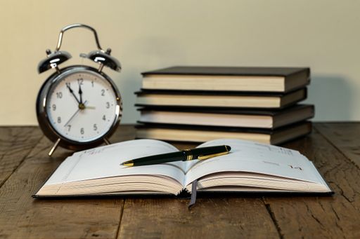 A closeup shot of a pen on a notebook, books, and an alarm clock on a desk