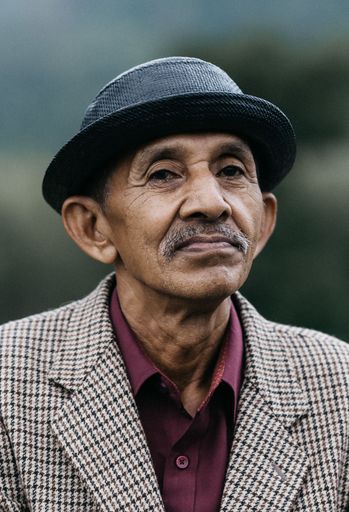 A vertical front closeup of an old man with a white mustache wearing a black hat and caged jacket on blurred background