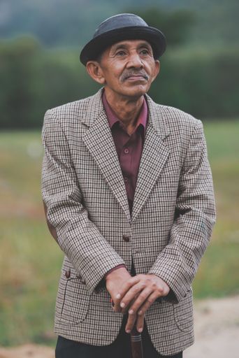 A vertical front shot of an old man with a white mustache wearing a black hat and caged jacket on blurred background