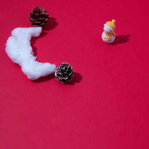 A top view of a small toy snowman in front of a cotton piece and pines on a red background