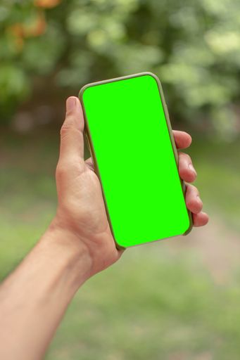 A vertical close-up shot of a man holding a smartphone in his hand with a green screen in a garden