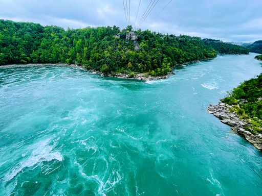 A high-angle closeup of Niagara Whirlpool with ropeway lines flowing water, green trees on both sides, and cloudy sky background