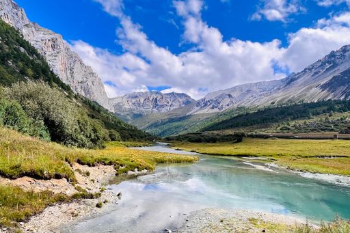 A long exposure shot of a clear lake with green hills and rocky mountains in the background.