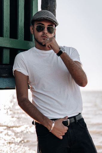 A vertical shot of a young sexy male in a white shirt, sunglasses, and newsboy cap posing on a beach