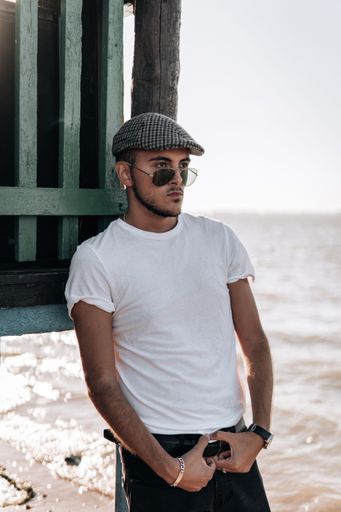A vertical shot of a young sexy male in a white shirt, sunglasses, and newsboy cap posing on a beach