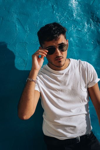 A vertical shot of a young sexy attractive male posing in sunglasses near a blue wall
