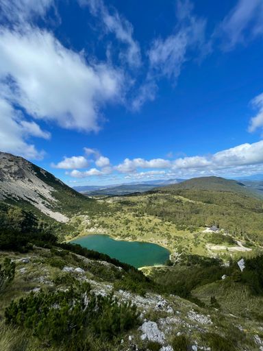 A vertical shot of a lake on a slope of green mountain under a cloudy blue sky