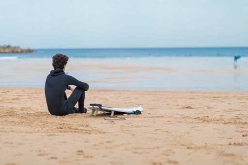 A curly-haired surfer sitting on the beach sand and looking at the seascape captured from behind