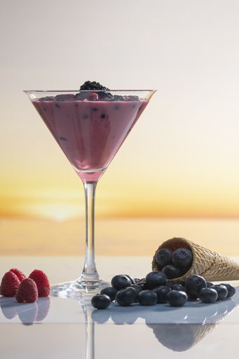 A vertical shot of Martini glass with fruit juice and blackberries surrounded by raspberries and blueberries with an ice cream cone on a glass table
