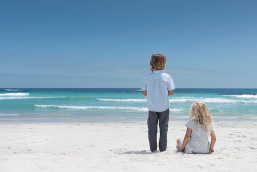 A back view of a boy standing on the beach and a girl sitting next to him on a sunny day